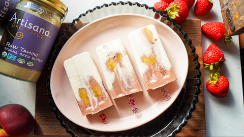 3 colorful strawberry peach popsicles with white chocolate coconut drizzle on them. A jar of tahini is off to the side.