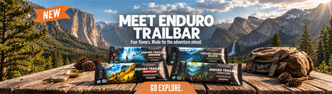 Four Enduro Trailbars are displayed on a rustic wooden table in a mountain landscape. The bars feature colorful scenic labels, and a brown backpack sits behind them.