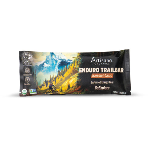 Artisana Organics Enduro Trailbar snack bar in a mountain landscape package design, featuring a cyclist on a trail with a snowy peak in the background. The package is black with yellow, green, and blue artwork, and text indicating 'Hazelnut-Cacao' and 'Sustained Energy Fuel'.