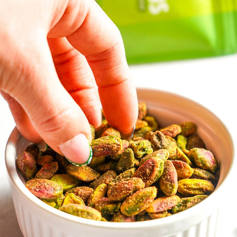 Hand holding a green pistachio over a bowl of colorful pistachios with a blurred green container in the background.