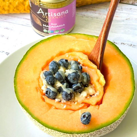 Half of a cantaloupe with macadamia butter and blueberries on a plate, next to a jar of Artisana Organics nut butter.