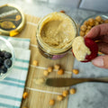 Person dipping a strawberry into a jar of macadamia cashew butter on a wooden mat with macadamia nuts and a bowl of blueberries.
